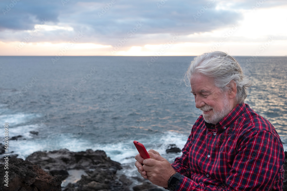Smiling senior man with white beard sitting on the cliff at the beach with horizon over water. Active happy retired people using phone