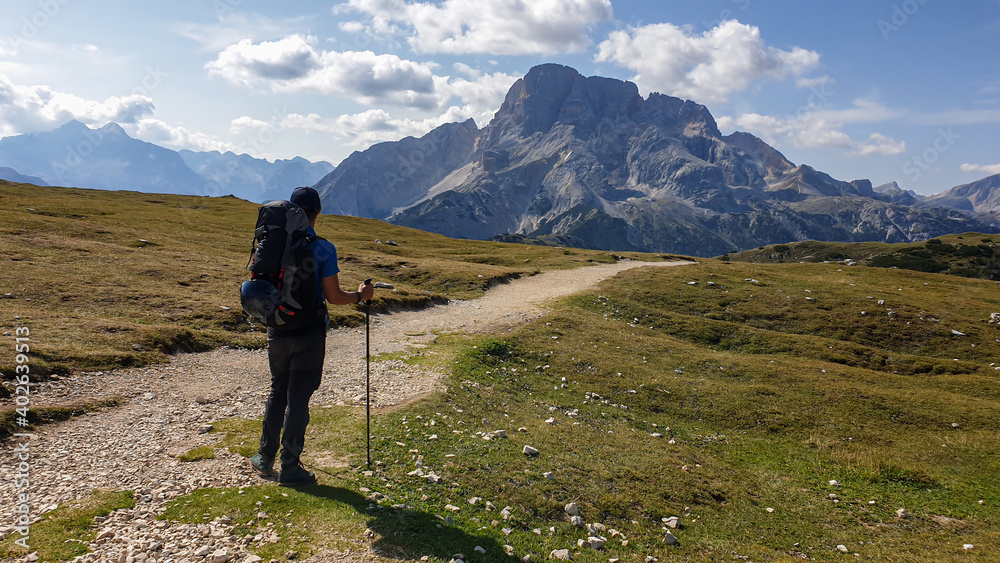 A man with hiking backpack hiking along a gravelled road in Italian Dolomites. There is a massive mountain chain in front of him, He is enjoying the trek. High Alpine plateau with lush green meadow. A man with hiking backpack hiking along a gravelled road in Italian Dolomites. There is a massive mountain chain in front of him, He is enjoying the trek. High Alpine plateau with lush green meadow.