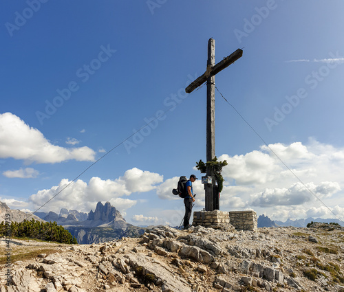 A man with a big hiking backpack standing under a massive wooden cross at the top of  Strudelkopf in Italian Dolomites. Drei Zinnen in the back. The man enjoys the view. High Alpine landscape. Freedom photo