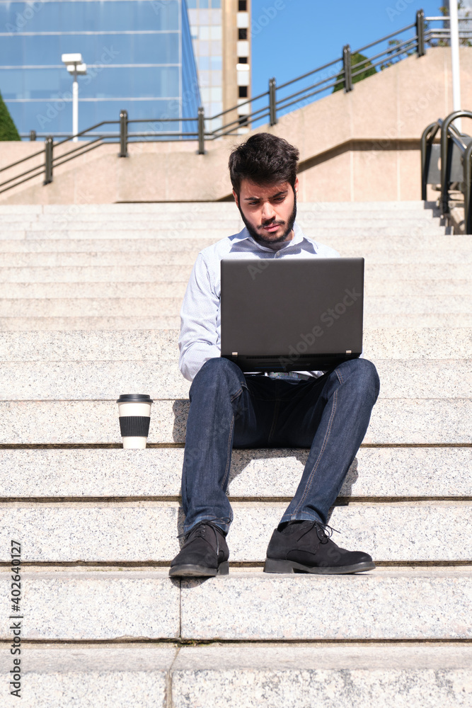Young business man using his laptop, with a coffee cup, sitting on stairs. Businessman in a urban street of a city.
