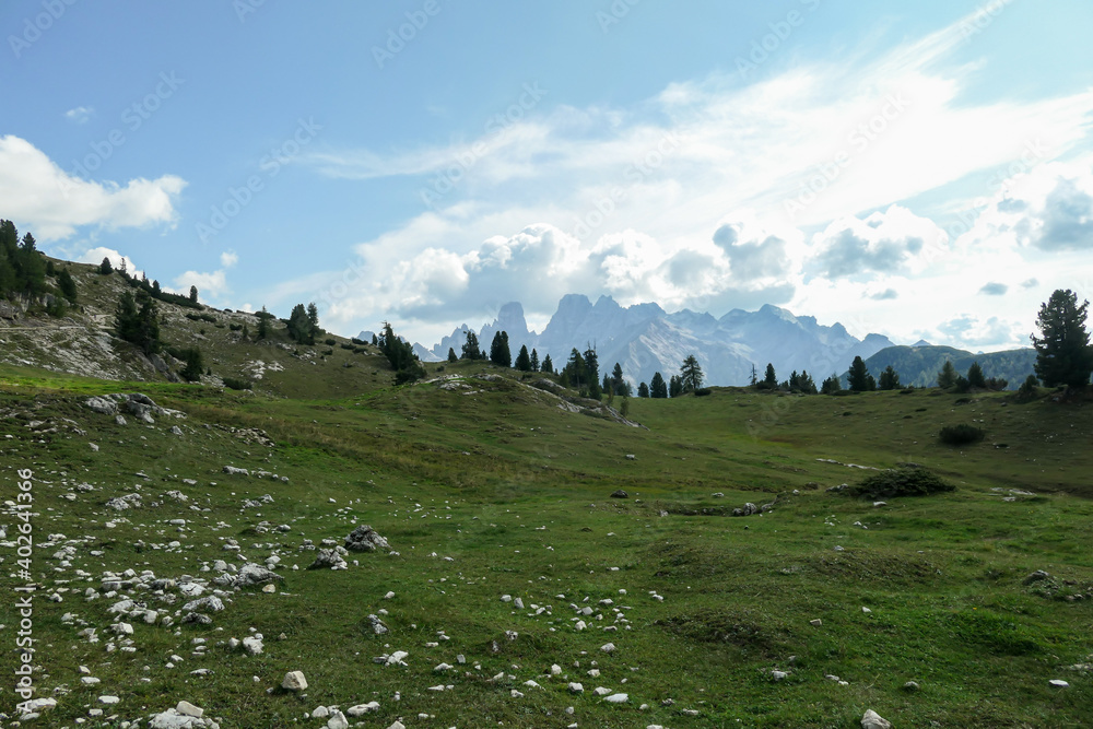 A panoramic view on the high Italian Dolomites, hiding in the clouds. Sunny day. A few clouds above the high peaks. Lush green plateau around with some pebbles and stones. Few trees growing in between
