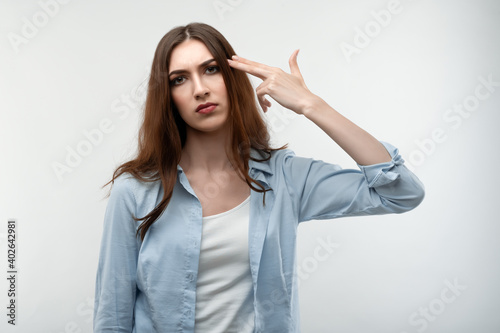 Unhappy caucasian Young woman with long chestnut hair, dressed in casual clothes, imitates gun shoot makes suicide gesture keeps two fingers on temples, has puzzled expression. Human emotions concept.