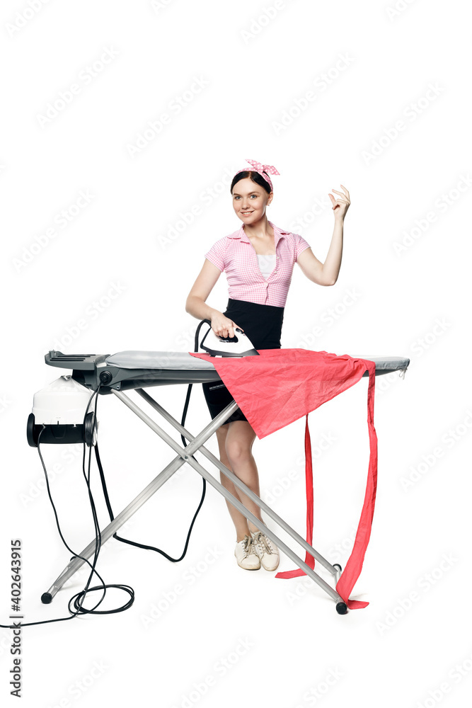 Fototapeta premium Snaps fingers. A beautiful girl dressed in pin-up style is ironing clothes looks at the camera and smiles isolated on a white background.