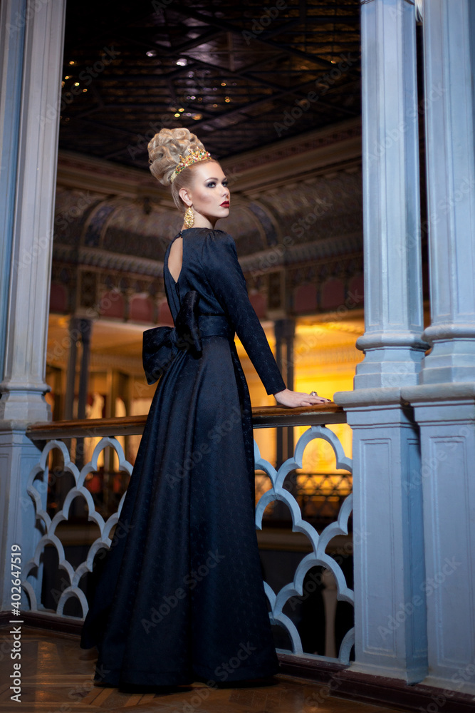 Gorgeous young blonde model, dressed in a long black dress with bow at the back, elegant hairstyle, crown and earrings.