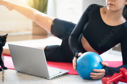 Fit young woman exercising while watching video on laptop. sporty woman is holding a pilates ball and lying on the floor with cat. online training. Sport and relaxation concept