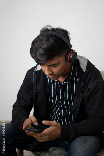 indian teenager boy black jacket with headphone sitting on floor at home. handsome guy looking at smartphone sctreen.