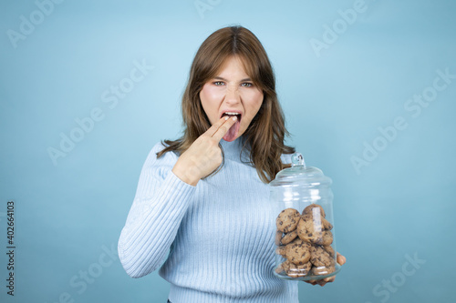 Young beautiful woman holding chocolate chips cookies jar over isolated blue background disgusted with her hand inside her mouth photo