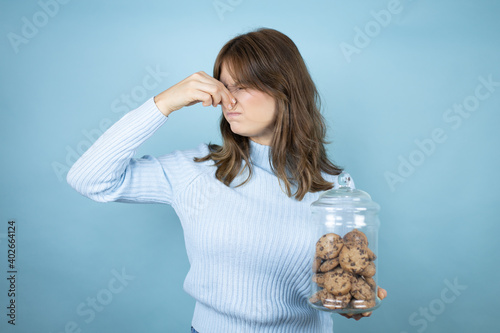 Young beautiful woman holding chocolate chips cookies jar over isolated blue background smelling something stinky and disgusting, intolerable smell, holding breath with fingers on nose photo