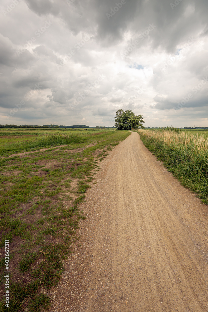 Dramatic cloudy sky over a winding and seemingly endless gravel road in a Dutch peat meadow area in the province of North Brabant.. The photo was taken in the beginning of the summer season. Dramatic cloudy sky over a winding and seemingly endless gravel road in a Dutch peat meadow area in the province of North Brabant.. The photo was taken in the beginning of the summer season.