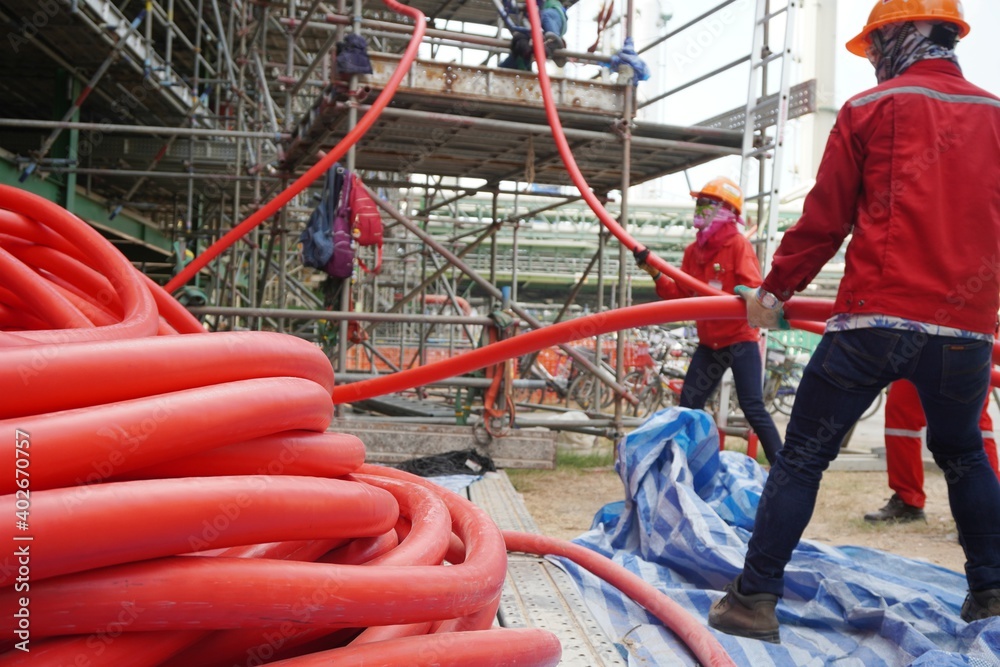 Large red power cord placed on the scaffolding floor and workers pulling high voltage cable line in chemical plants, power plants, oil & gas industry or onshore. Large red power cord placed on the scaffolding floor and workers pulling high voltage cable line in chemical plants, power plants, oil & gas industry or onshore.