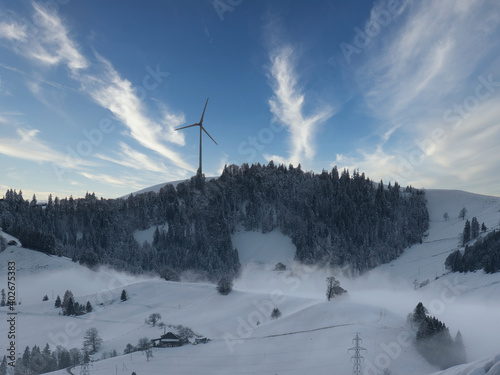 Schneebedeckte Landschaft mit Hügeln, Wald und Windkraftanlage an einem kalten Wintertag in der Gemeinde Schwarzenberg (Schweiz)