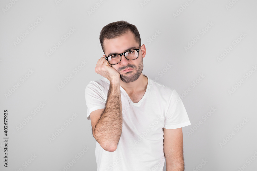 Studio shot of an annoyed annoyed handsome guy in black glasses who touches his face and bends over, looks out from under his eyes, hates talking to a boring person, stands against a gray background Studio shot of an annoyed annoyed handsome guy in black glasses who touches his face and bends over, looks out from under his eyes, hates talking to a boring person, stands against a gray background