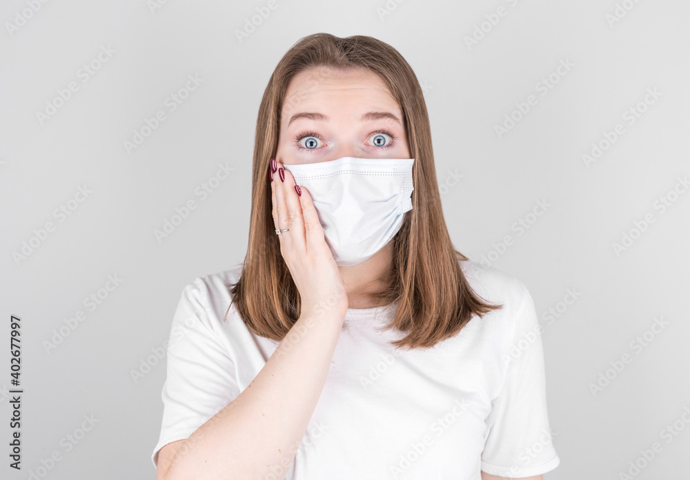 Young female doctor in a medical mask looks into the camera with surprise, holding her face with her hand. after contracting COVID 19, flu and seasonal colds.