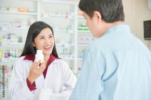Smiling asian professional young pharmacist showing medicine bottle to customer at drugstore.