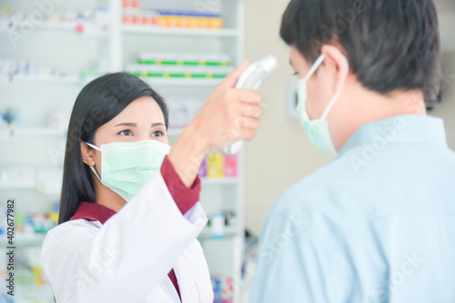 Asian professional pharmacist wearing mask checking customer temperature by digital thermometer at drugstore.