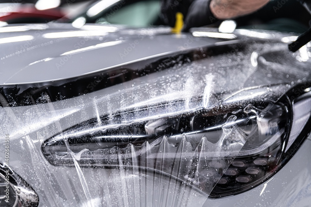Car detailing studio worker applying protective ppf foil film on car body and lamps