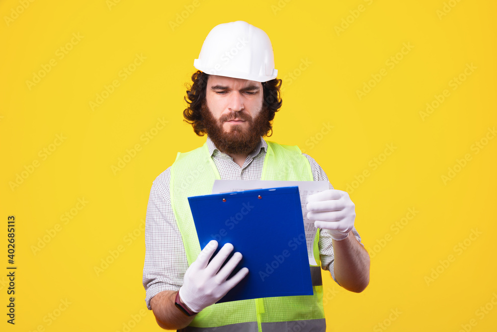 An handsome bearded engineer is looking confused at his work pappers near a yellow wall . An handsome bearded engineer is looking confused at his work pappers near a yellow wall .