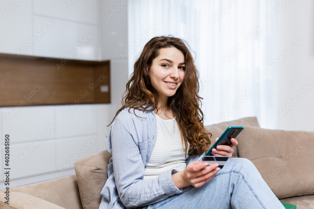 Portrait happy female shopper using instant easy mobile payments making purchase in online store young woman customer holding credit card and smartphone sitting on couch at home Portrait happy female shopper using instant easy mobile payments making purchase in online store young woman customer holding credit card and smartphone sitting on couch at home