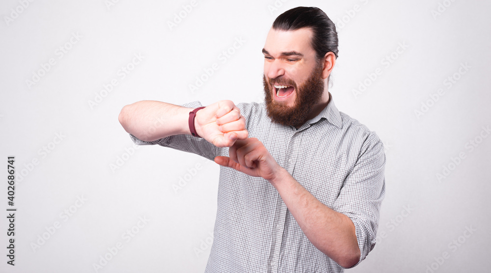 A bearded young man is looking stressed at his watch being afraid that he is late near a white wall . A bearded young man is looking stressed at his watch being afraid that he is late near a white wall .