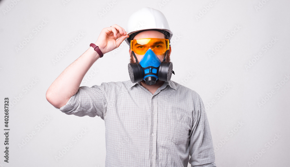 Obraz premium Young bearded engineer wearing a helmet, protective glasses and a respirator is looking at the camera near a white wall .