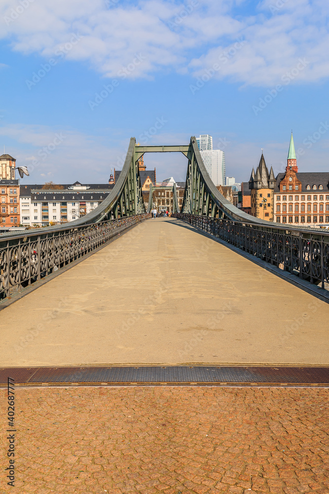 Obraz premium Historic footbridge over the Main in Frankfurt by day. Houses of the old town in the background with sunshine and a few clouds. Bridge with iron terrain
