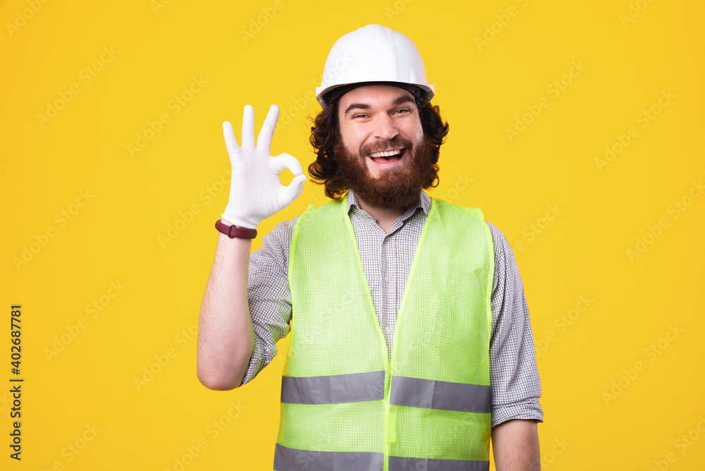 An nice bearded young architect is looking cheerful at the camera showing the ok sign near a yellow wall .