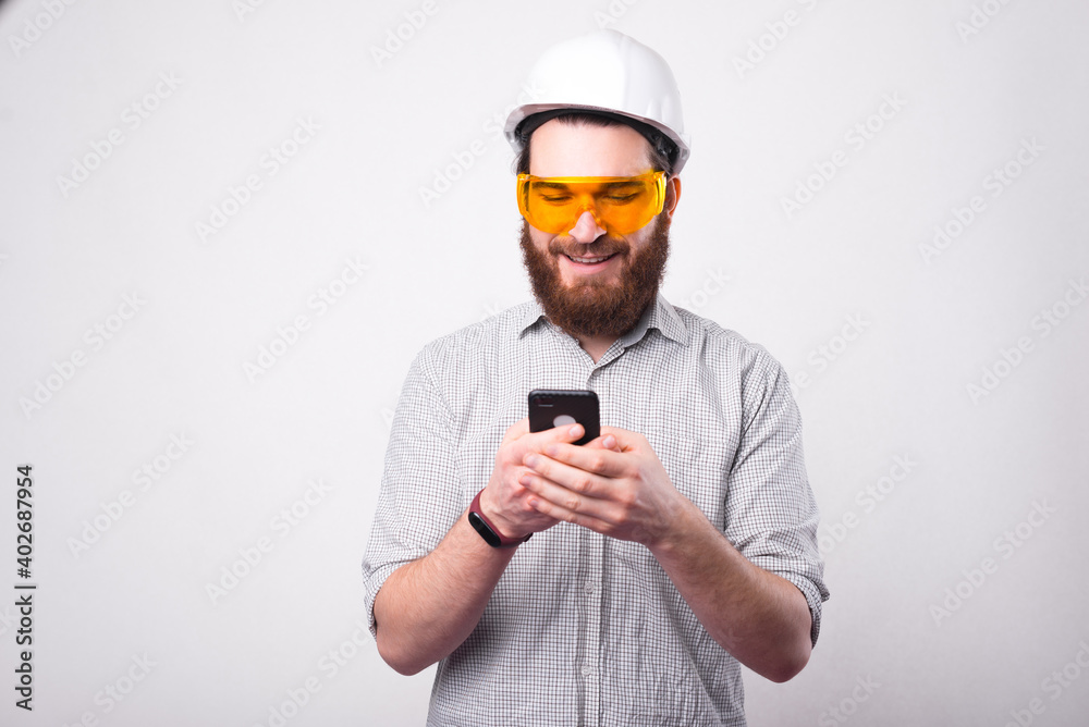 A young architect is texting happy on his phone wearing a pair of protective glasses and a hemlet near a white wall .