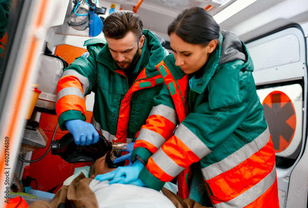Two paramedics are resuscitating a senior lying on a gurney in an ambulance by performing chest compressions and connecting to a ventilator. Two paramedics are resuscitating a senior lying on a gurney in an ambulance by performing chest compressions and connecting to a ventilator.