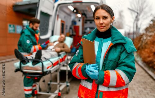 A woman paramedic in uniform stands with a patient card in front of an ambulance and her colleague standing near a patient's gurney.