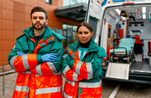 Two paramedics in uniforms stand with their arms crossed in front of a clinic and a modern ambulance.
