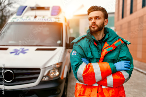 An adult handsome male paramedic stands with his arms crossed in front of an ambulance near the clinic. © Valerii Apetroaiei