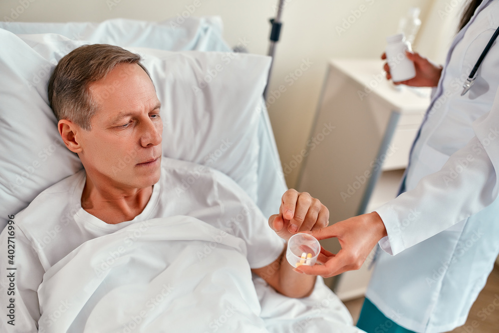 A beautiful young nurse gives a mature male patient a medicine to drink, who lies on a bed in a modern hospital ward. A beautiful young nurse gives a mature male patient a medicine to drink, who lies on a bed in a modern hospital ward.