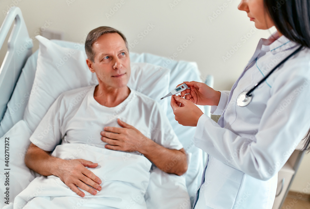 A beautiful female doctor measures the temperature of a mature male patient who is undergoing rehabilitation in a modern hospital ward. A beautiful female doctor measures the temperature of a mature male patient who is undergoing rehabilitation in a modern hospital ward.
