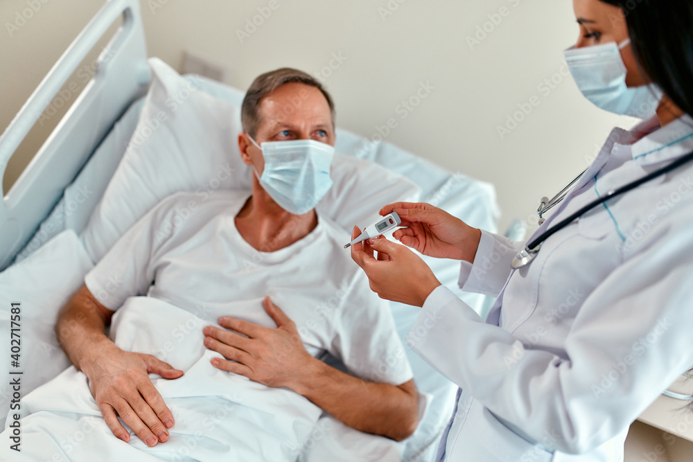 A female doctor in a protective medical mask measures the temperature of a mature male patient who is undergoing rehabilitation in a modern hospital ward during the coronavirus or covid-19 epidemic. A female doctor in a protective medical mask measures the temperature of a mature male patient who is undergoing rehabilitation in a modern hospital ward during the coronavirus or covid-19 epidemic.