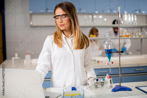 Female researcher in white lab coat and protective mask using laptop while working in the lab