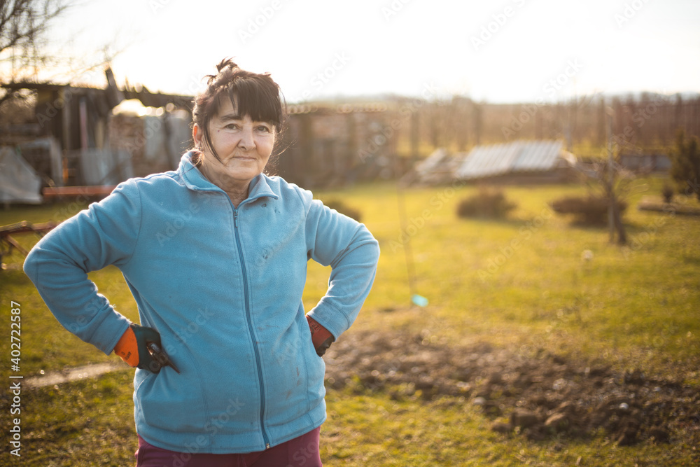 Obraz premium Portrait of beautiful happy older woman in her garden standing and smiling outside during sunset. Wind Blows Her Hair. Beautiful Sunset Day