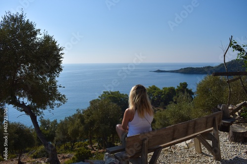 Young blonde woman sitting on a bench on top of a hill and watching sea