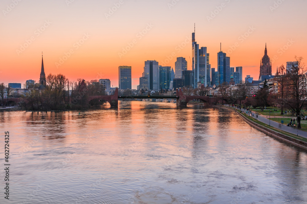 Frankfurt skyline in the evening. Sunset over the river Main from the city center. Building of the financial and business district. Park along the bank with trees and meadow