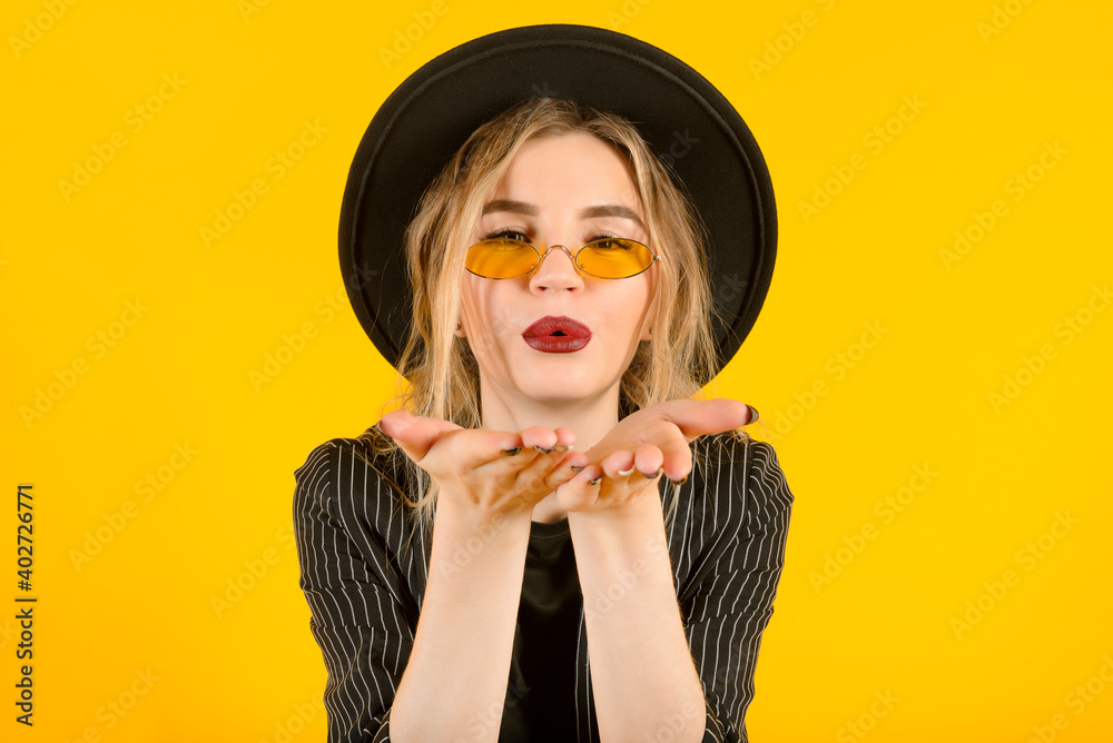 stylish young blonde woman in a black suit, hat and yellow glasses on a yellow background blows a kiss stylish young blonde woman in a black suit, hat and yellow glasses on a yellow background blows a kiss