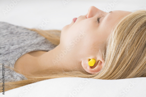 Young adult blonde woman sleeping with yellow earplug into her ear in bed. Head on pillow. Very well asleep in silence. Side view. Closeup.