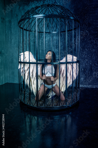 Angel girl is locked in a cage and kneels illuminated by cold blue light