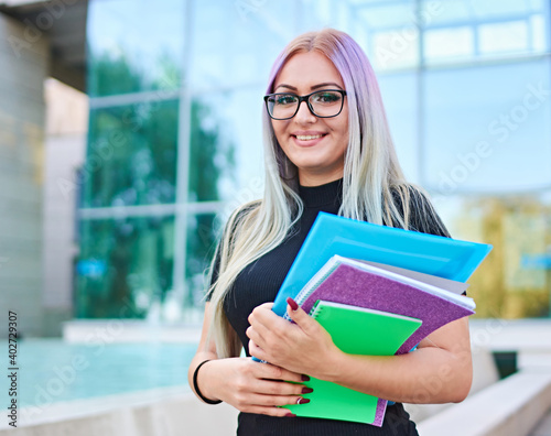 Young happy student girl standing in front of a campus building and looking into camera - Smart millennial woman with glasses holding booklets - Freshman with colorful hair smiling after an exam