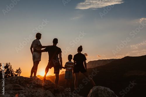Friendly embracing family with two kids, a son and daughter, stand on top of mountain and watch colorful sunrise and blue sky