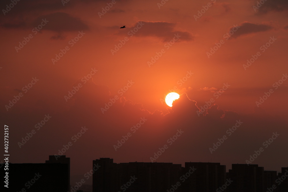 Sunset over the city. Sunset city scenery with sun. City silhouette against the sky on a sunset. 
Sun hiding behind the cloud.