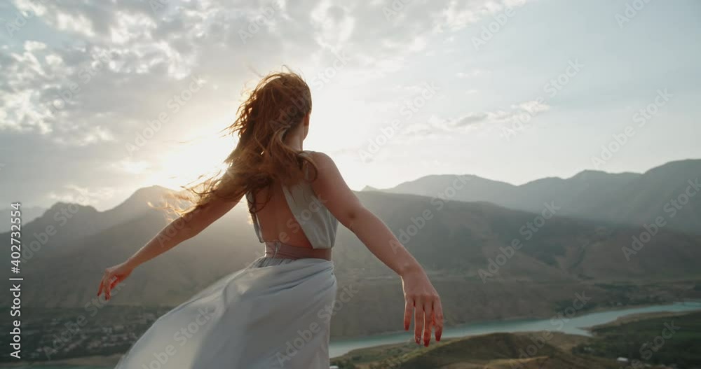 Woman in white dress standing on top of a mountain with raised hands while wind is blowing her dress and red hair - freedom, nature concept 4k footage