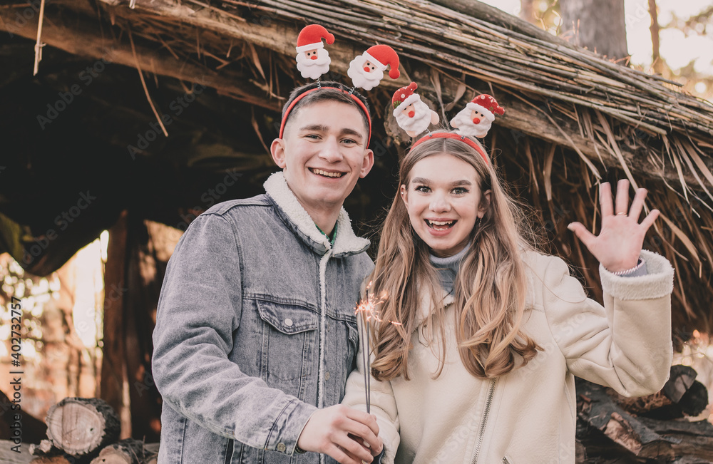A couple about their appearance, dressed in New Year's hoops on their heads, holds a sparkler in their hands and smiles against the background of a gazebo made of firewood