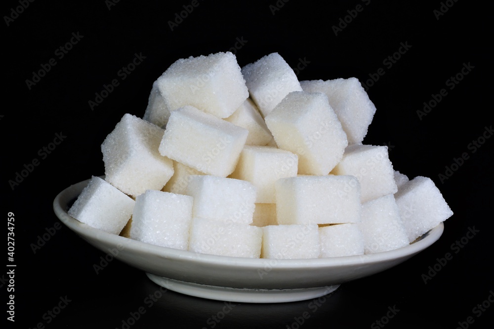 Sugar is white in a plate on a black background. It is used in food products and added to beverages. Sugar is white in a plate on a black background. It is used in food products and added to beverages.