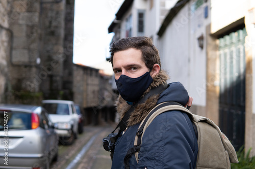 Photo of a young and attractive man wearing a face mask and a backpack and travelling with a photo camera. Doing tourism in an empty city