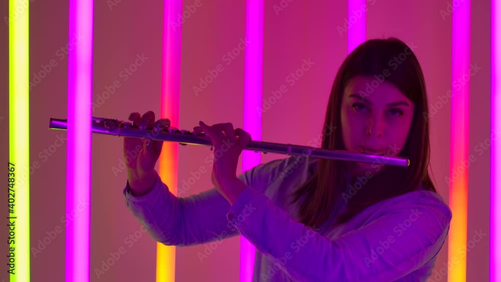 Portrait of young woman musician blowing into wind instrument flute. A female flutist performs in a dark studio against the backdrop of bright neon lights. Silhouette. Slow motion. Close up. Portrait of young woman musician blowing into wind instrument flute. A female flutist performs in a dark studio against the backdrop of bright neon lights. Silhouette. Slow motion. Close up.
