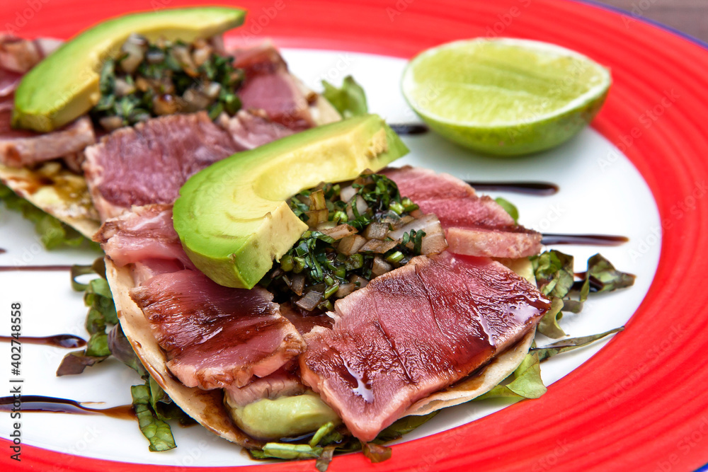 Mexican tostadas with shaved tuna, onion seasoned with soy sauce, slices of avocado and lemon, served on a red and white plate on a wooden background